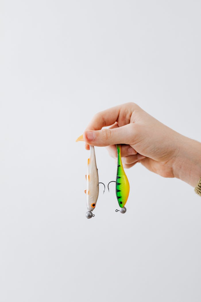 who-we-are An adult hand holding two colorful fishing lures against a neutral background.